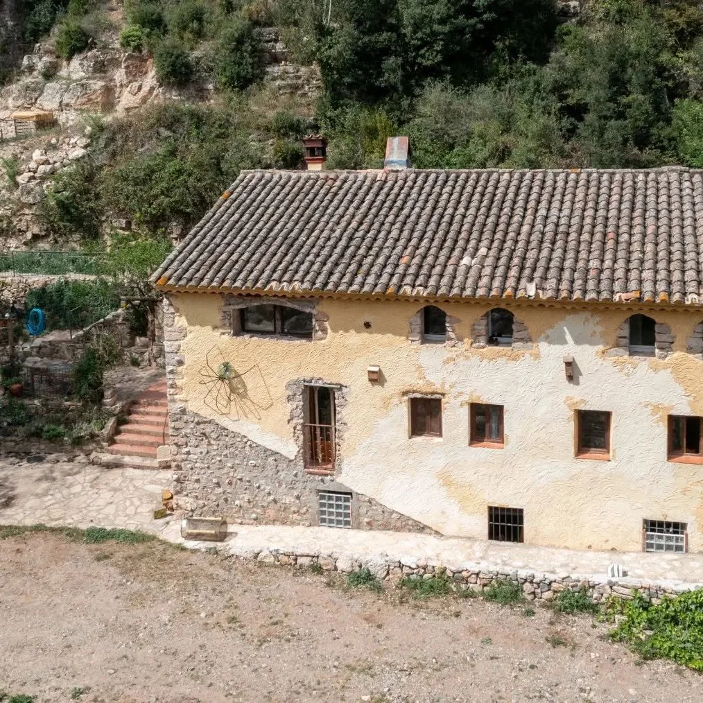 Façana de pedra i teulada de teules del Molí del Fort, al costat d’un vessant arbrat.